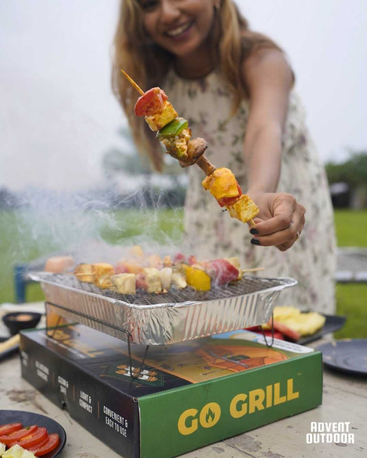 Smiling woman holding grilled paneer skewer over a lit Go Grill disposable BBQ on a picnic table — perfect for outdoor cooking and weekend gatherings.