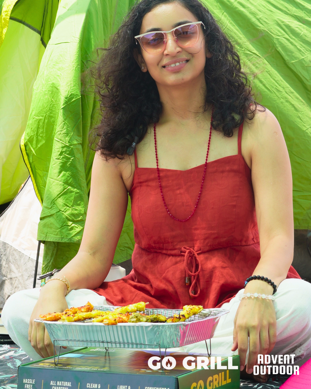 Woman enjoying outdoor camping with Go Grill disposable BBQ placed on its box, grilling food beside a green tent — ideal for easy travel cooking and picnics.