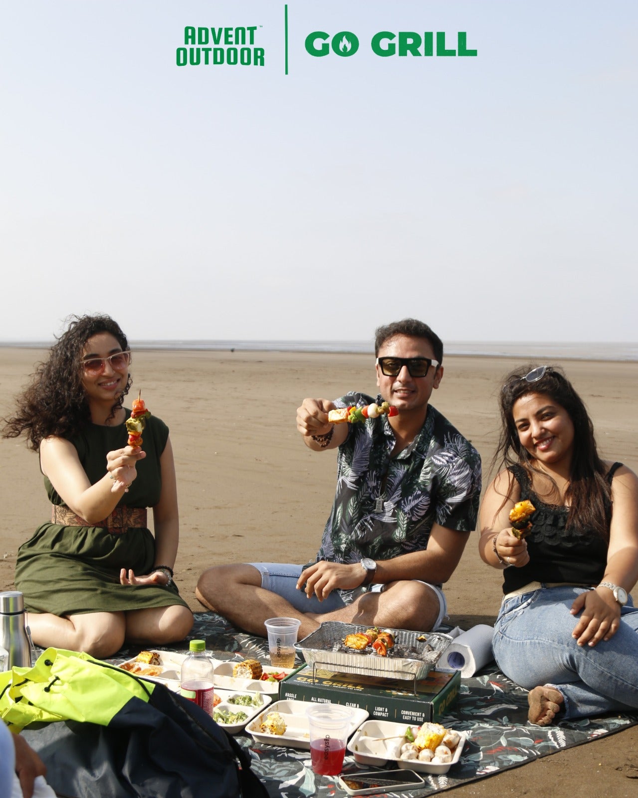 Group of friends enjoying grilled skewers with Go Grill disposable BBQ at a beach picnic, surrounded by food and drinks — perfect for travel-friendly outdoor grilling.