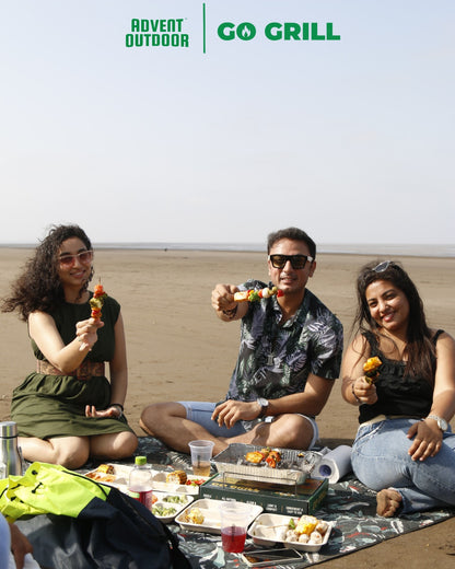 Group of friends enjoying grilled skewers with Go Grill disposable BBQ at a beach picnic, surrounded by food and drinks — perfect for travel-friendly outdoor grilling.